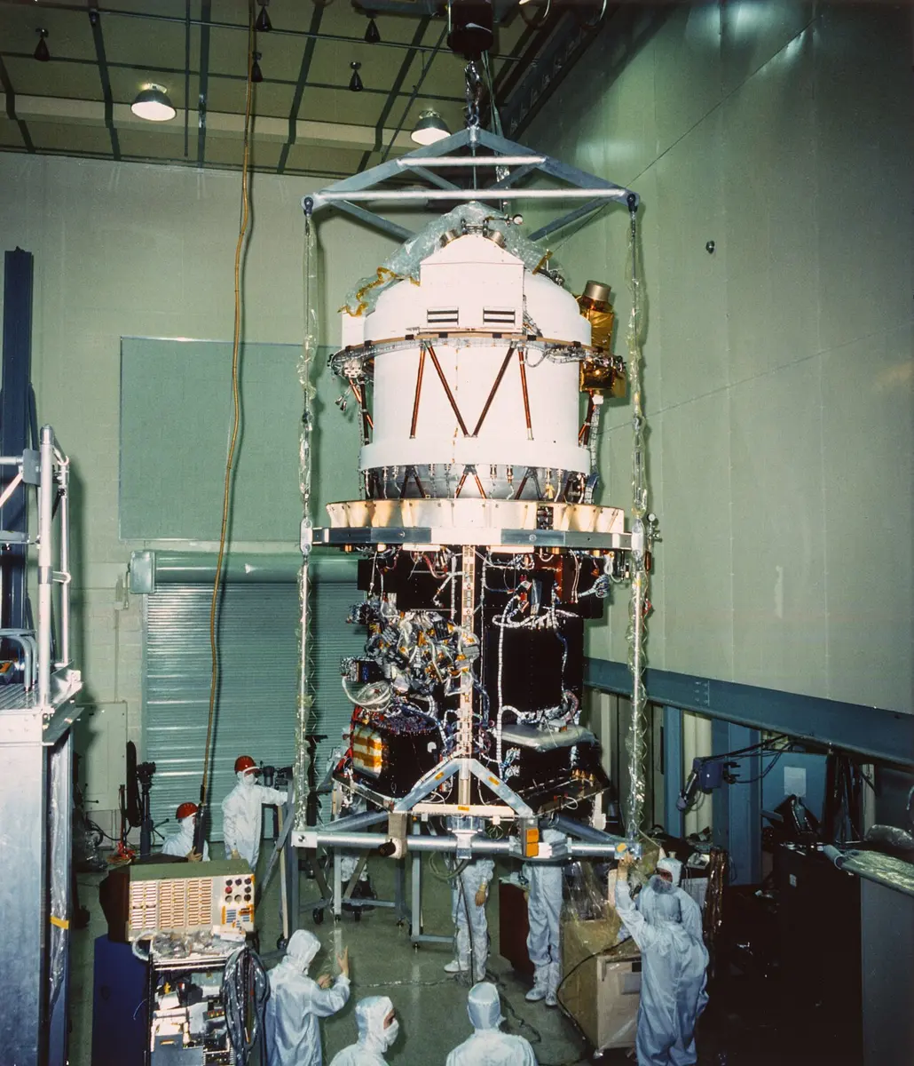 A tall white and silver spacecraft hanging from a crane inside a large clean room, with workers in white suits and caps around it