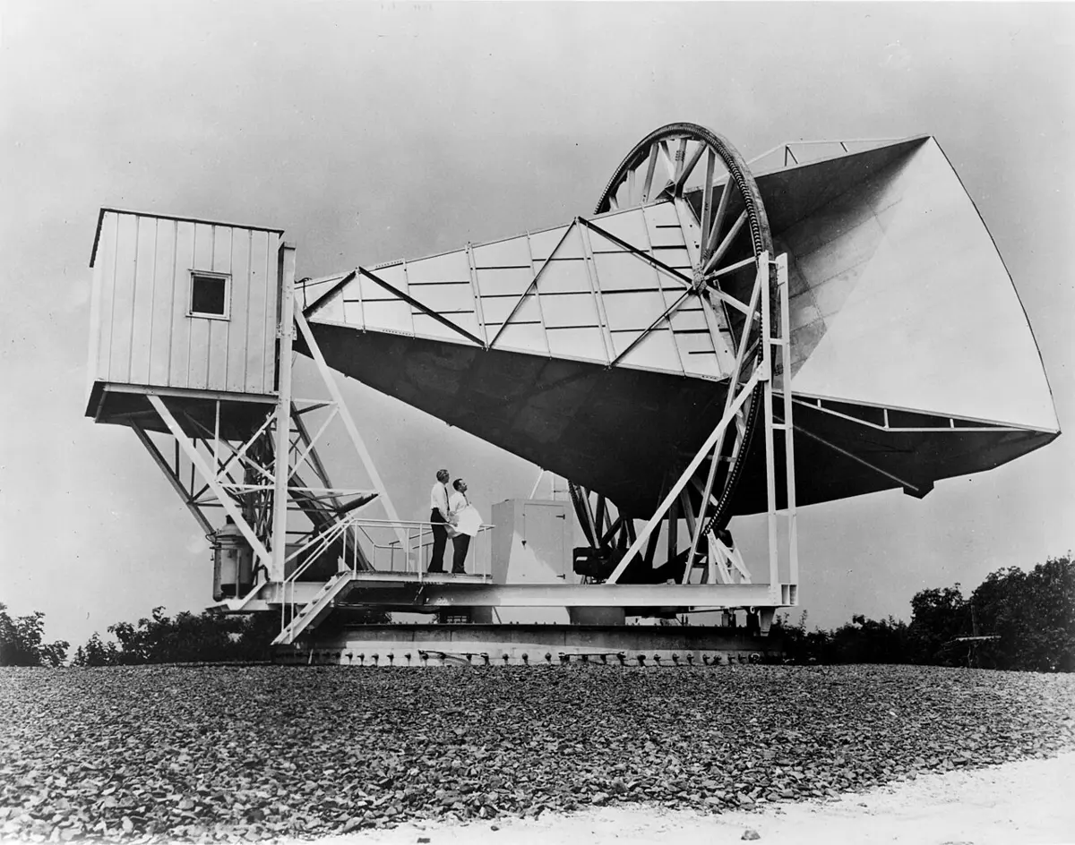 A black-and-white photo of a huge horn-shaped metal antenna, like a giant funnel lying on its side, mounted on a frame with wheels, with two men standing on it