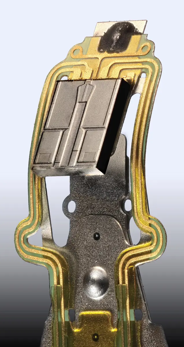A close-up of the tip of a hard disk arm: a small gray rectangular block on a flexible strip with gold-colored wires
