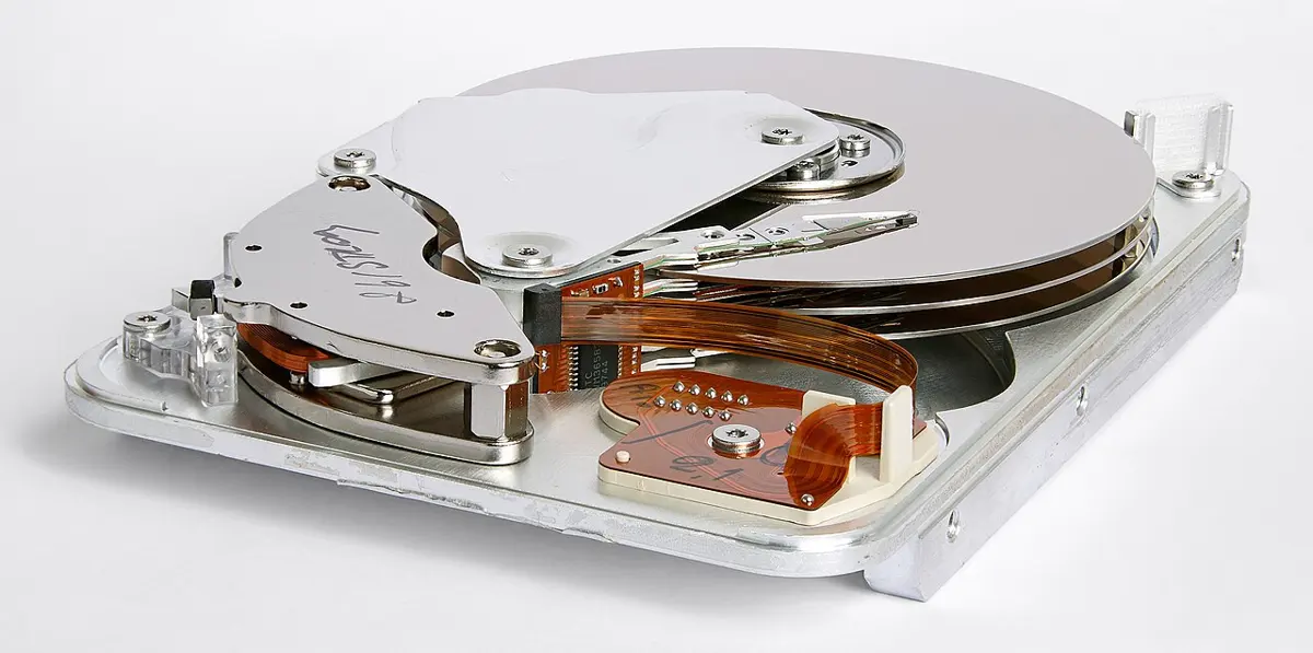An opened hard disk drive: a shiny silver disk, and a metal arm that reaches across it, with a thin flat strip of copper-colored wires attached