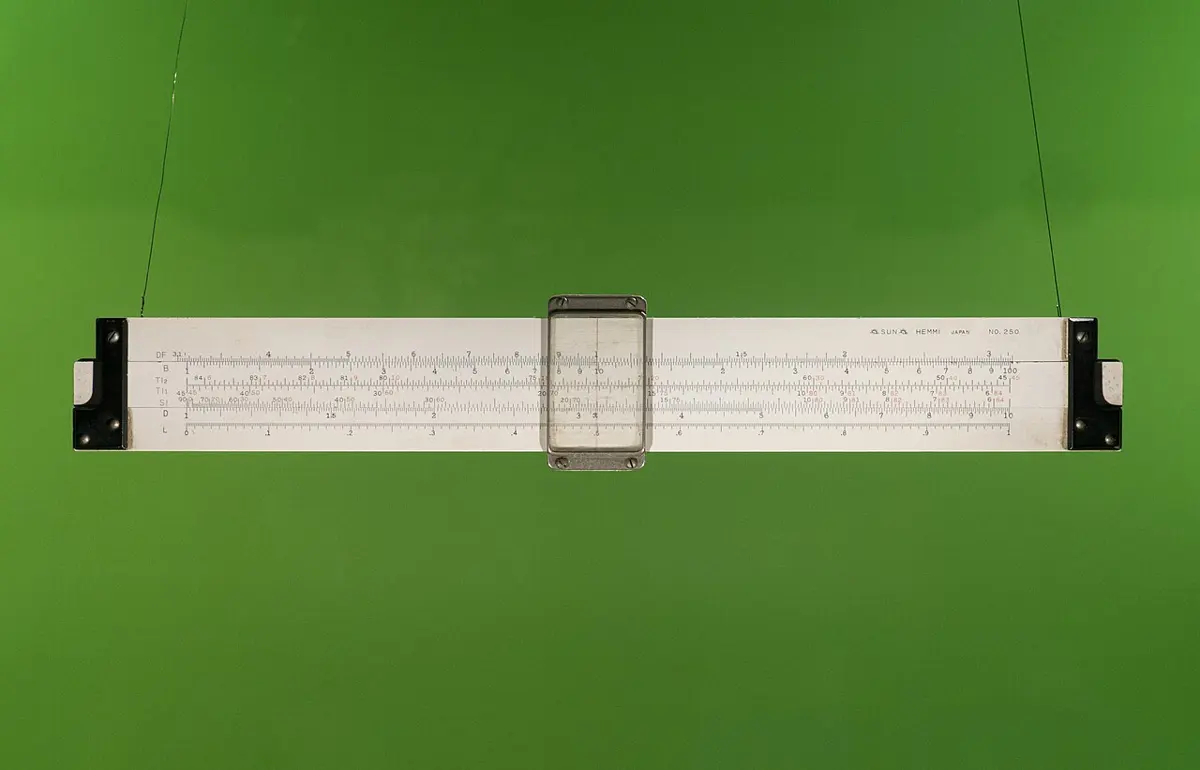 A long white slide rule, a ruler with a sliding middle strip and a clear window marked with fine scales, hanging against a green background