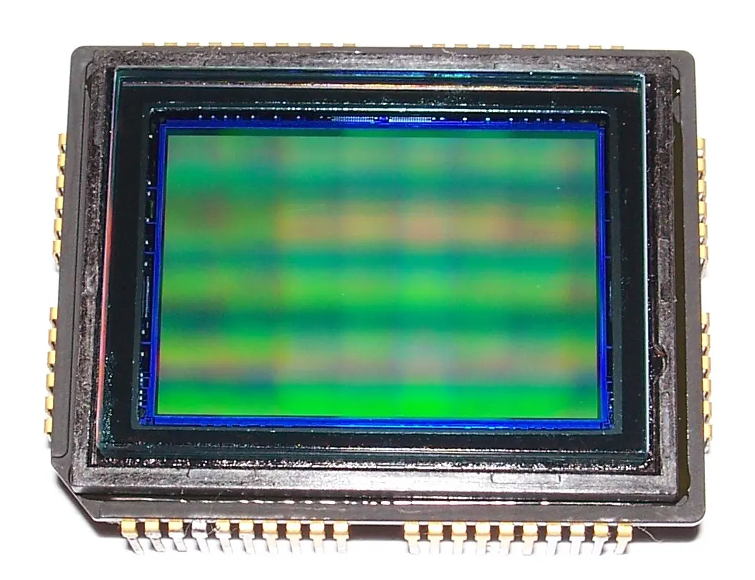 A rectangular image sensor in a black package with gold contact pins along its edges; the sensor surface in the middle shimmers green with faint bands