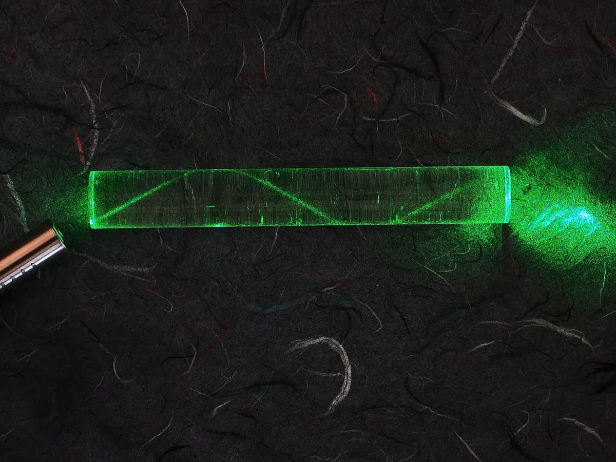 A short clear plastic rod lying on dark cloth, lit from its left end by a green laser pointer; inside it the beam zigzags from side to side along its length