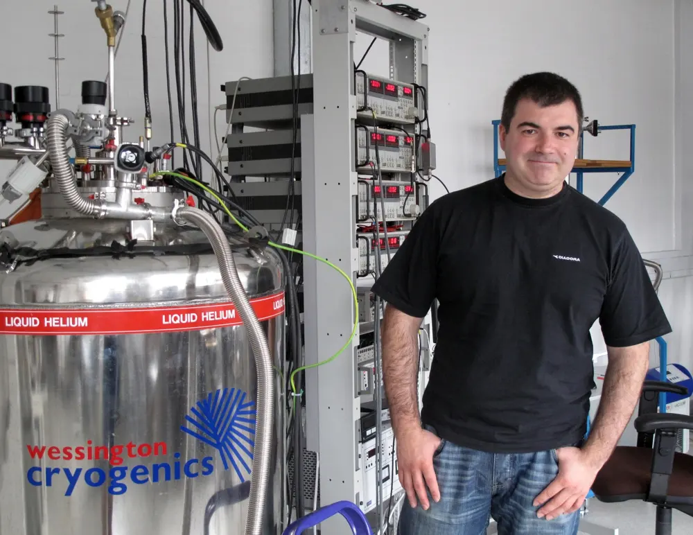A smiling man in a black T-shirt stands in a laboratory beside a large shiny metal tank labeled “liquid helium” and racks of electronic equipment with red number displays