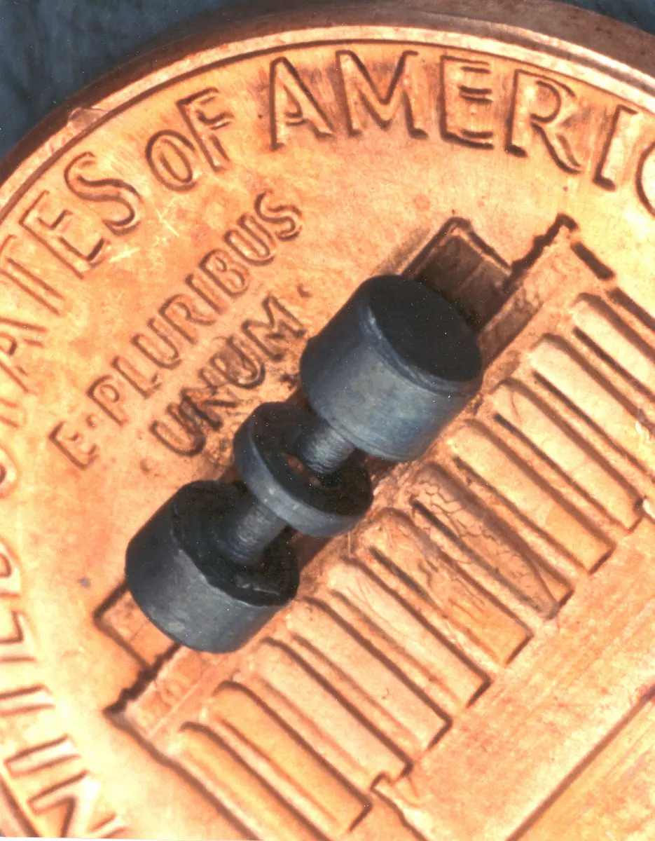A small dark metal part shaped like a short rod with two caps and a ring, lying on a copper coin with ‘E pluribus unum’ printed on it