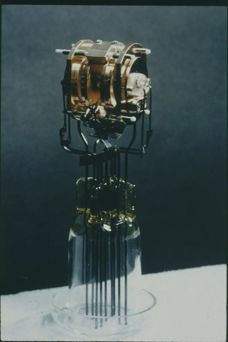 An old photograph of a small gold-colored metal cylinder made of rings, held up on thin wires above a glass base