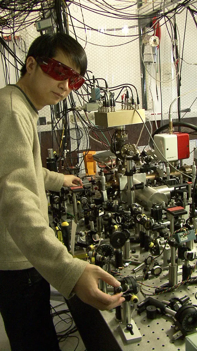 A young researcher in red safety glasses beside a table crowded with lenses, mirrors and cables, under a tangle of wires