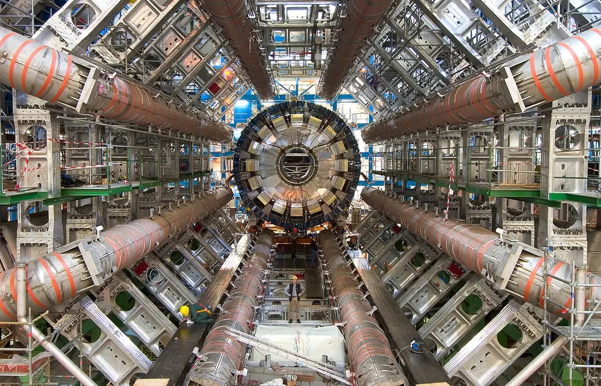 A huge underground hall: eight giant silver tubes with orange bands run away from the viewer in a ring round a big round machine in the middle; a man in a hard hat stands at the bottom, tiny beside them