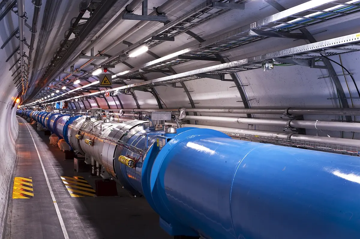 A long, curving concrete tunnel lit by lamps, with a row of large blue cylinders, the magnets, running along one side