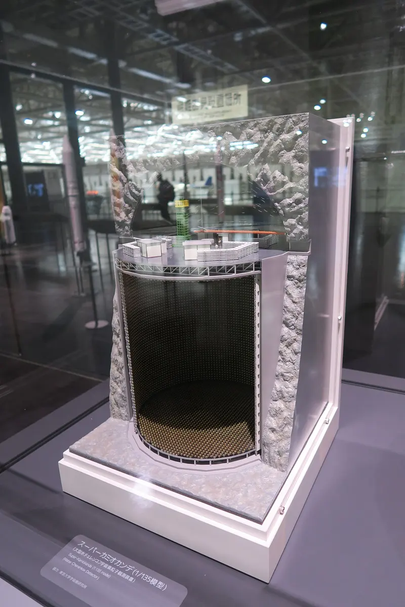 A museum model of a tall cylindrical tank cut open inside a block of gray rock, its inner walls covered in rows of small golden dots, with a platform and small buildings on its lid