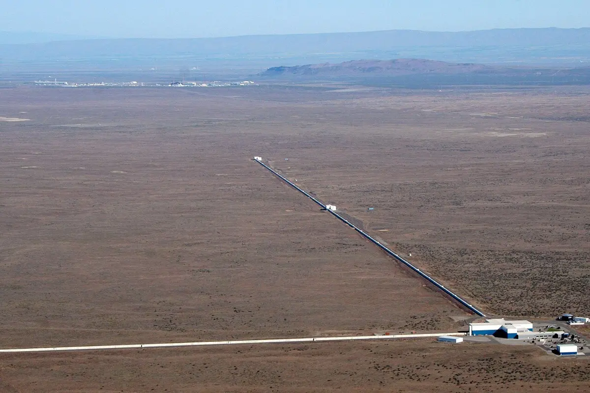 An aerial photograph of flat, dry brown land with two long, straight, narrow concrete tunnels meeting at a right angle at a group of buildings