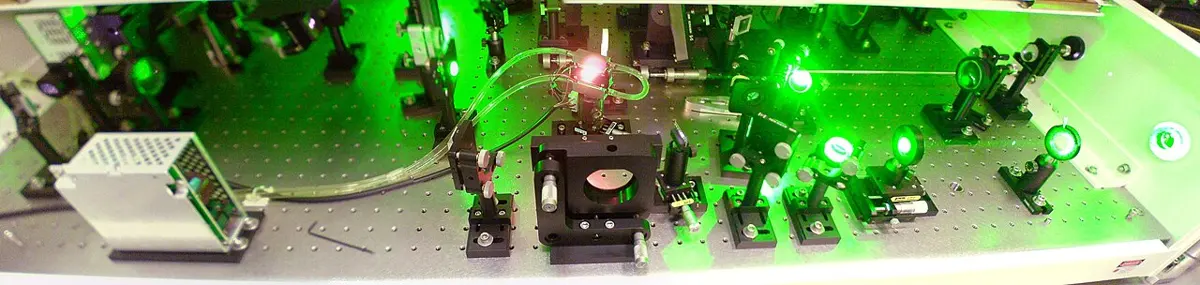 A long table covered with mirrors, lenses and metal mounts, lit by bright green laser light, with a glowing crystal in the middle