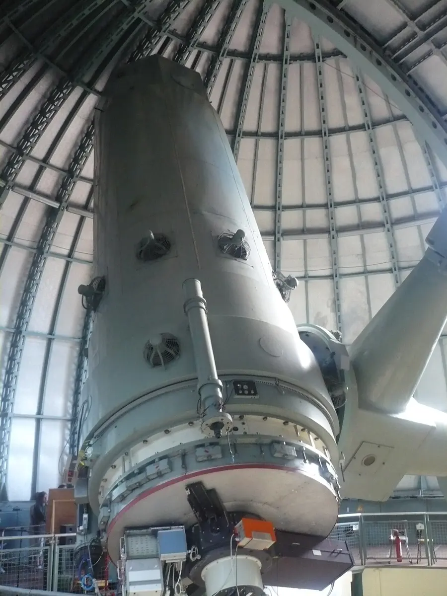 A huge gray cylindrical telescope tube on a heavy mount, pointing up inside a dome with a ribbed metal roof