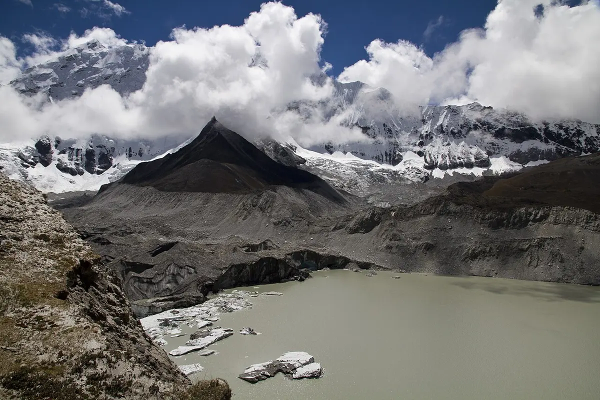 A gray-green lake with floating chunks of ice, ending at a wall of glacier ice covered in gray rock, with snowy mountains and clouds behind