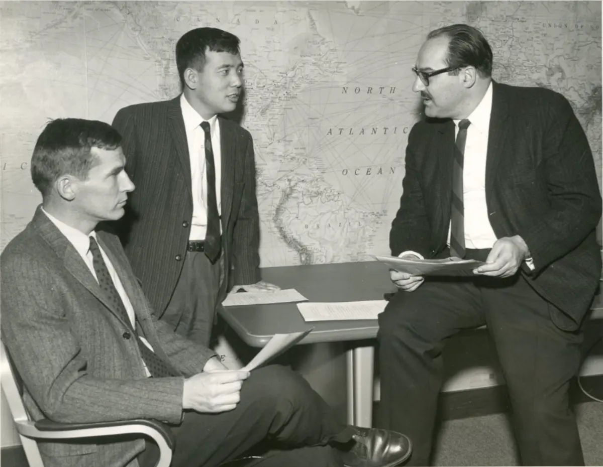 An old black and white photo of three men in suits and ties talking in an office, in front of a large wall map of the Atlantic Ocean