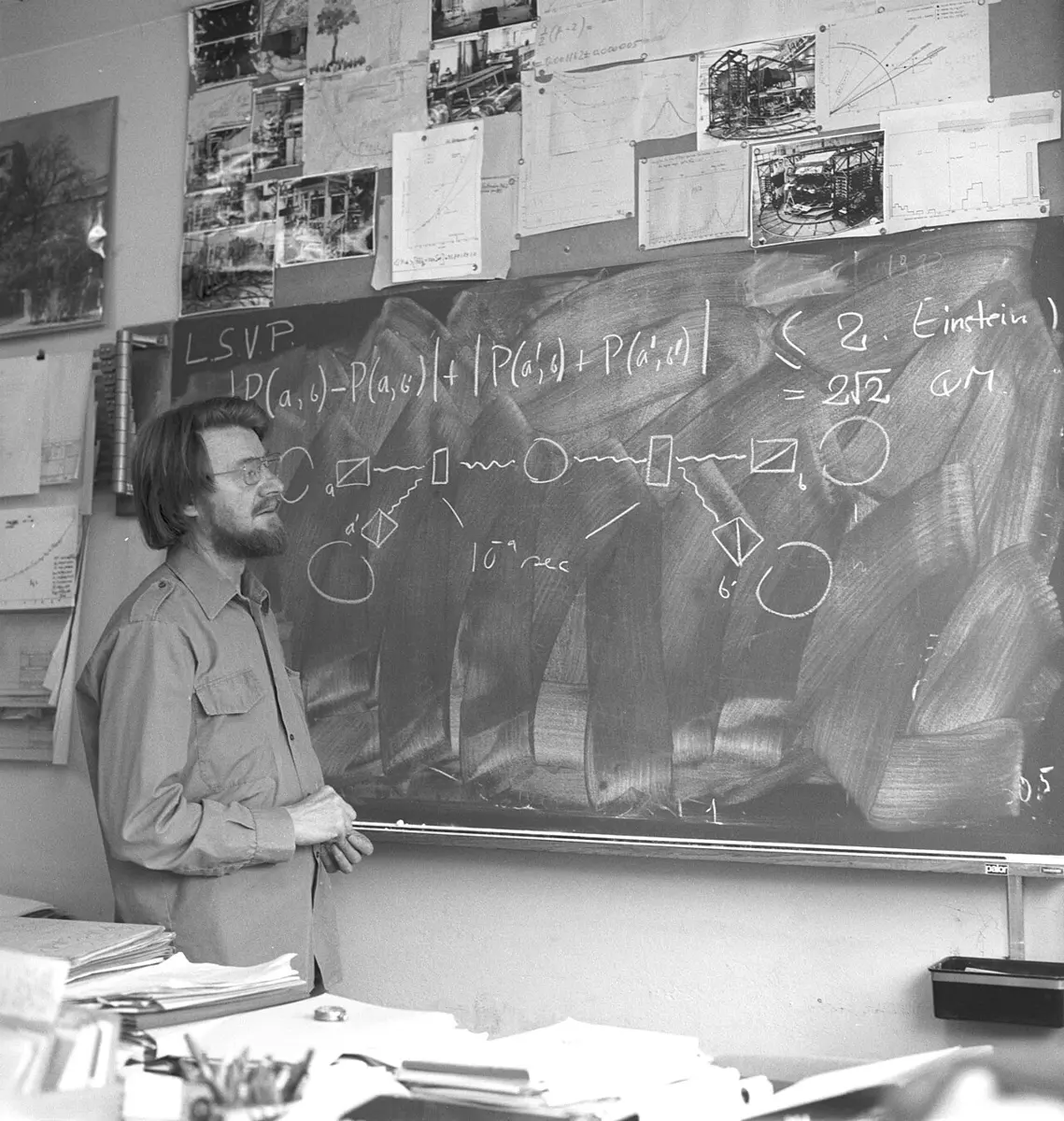 Black and white photo of a bearded man with glasses and long hair standing at a blackboard with a formula and a drawing of a source, two switches and four detectors