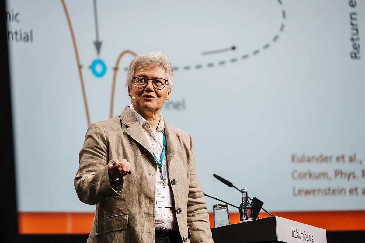 Anne L’Huillier, with short gray hair and glasses, speaking on a stage, in front of a large screen showing a diagram of an electron leaving an atom and coming back