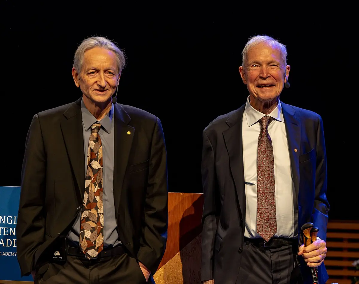 Two smiling elderly men in dark suits and patterned ties standing side by side on a stage with a dark background; the one on the right holds a walking stick