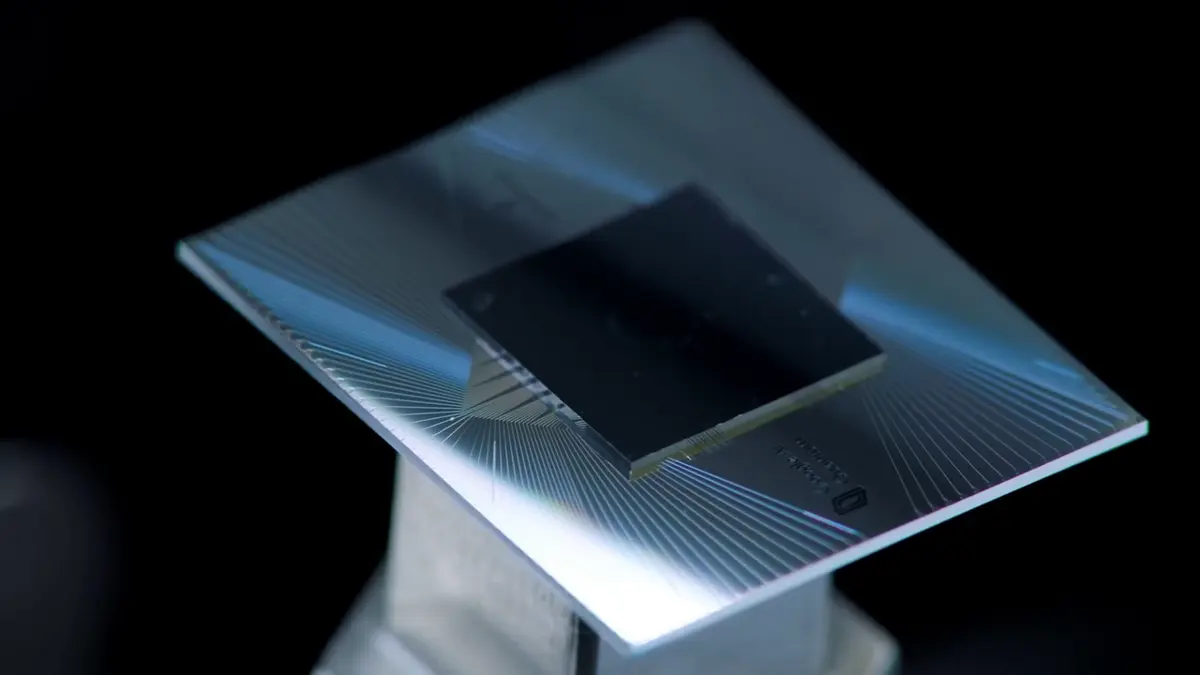 A small dark square chip mounted on a larger shiny silver square, with fine lines fanning out from the chip to the edges, on a metal stand against a black background
