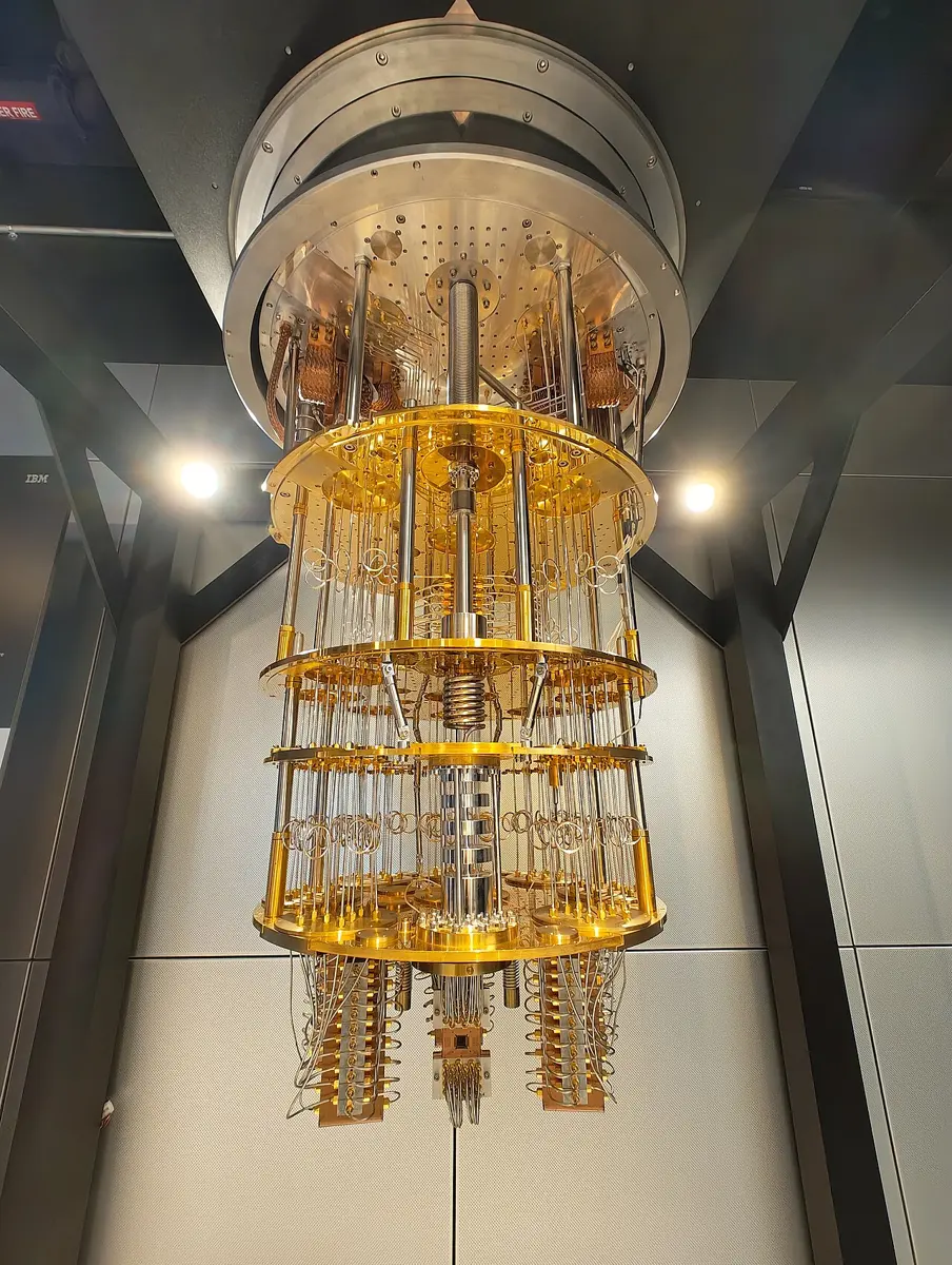A tall cylinder of golden metal plates connected by rods and coiled cables, hanging from the ceiling in a lit glass case