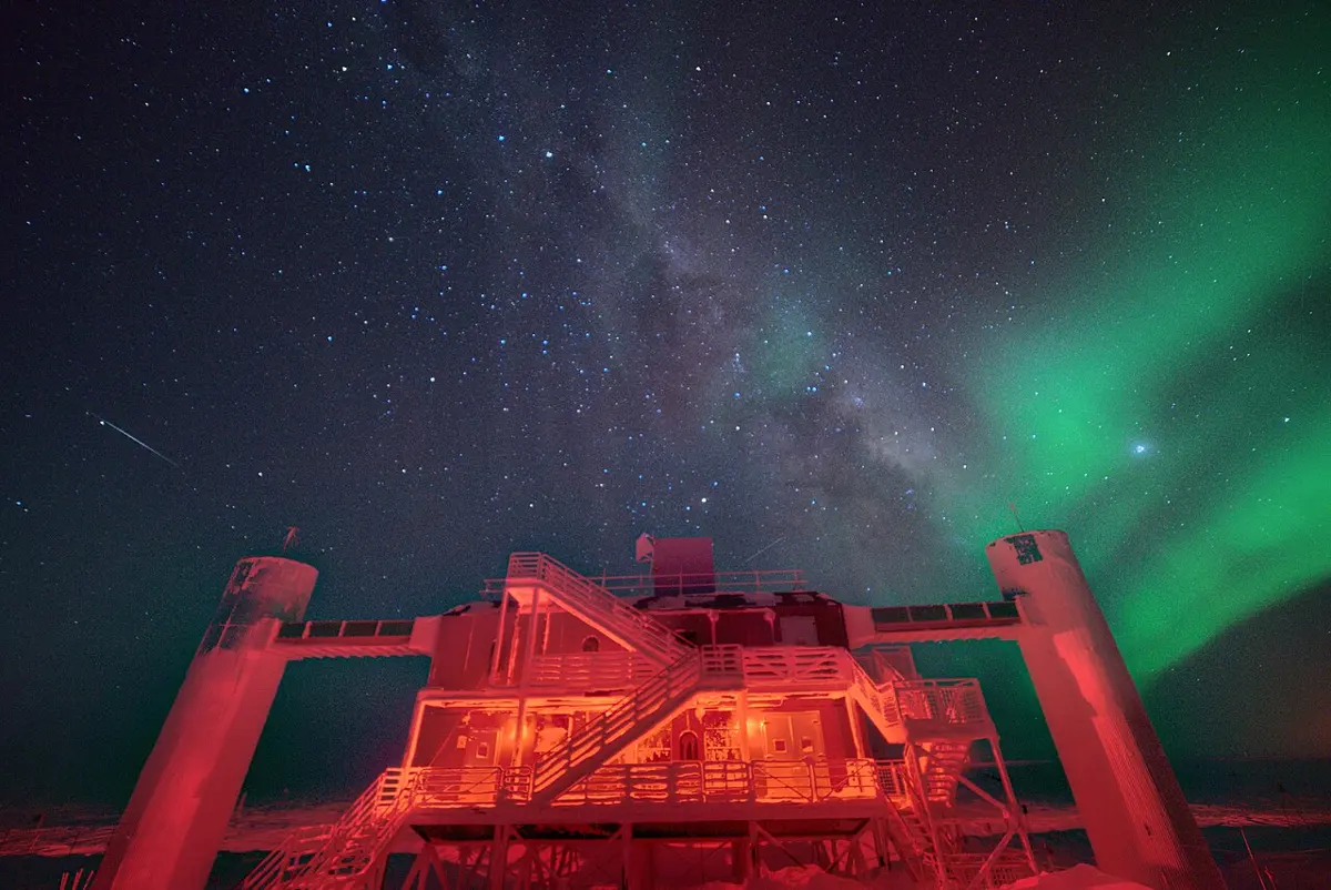 A two-storey building with outside stairs, standing between two towers on the snow, lit red at night, under a starry sky with the Milky Way and a green aurora